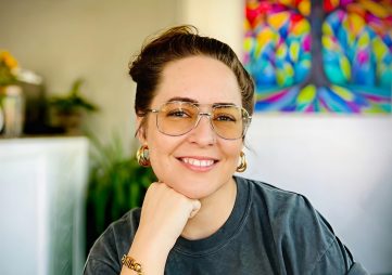 White woman with brown hair in a top bun, golden aviator glasses, and a dark great t-shirt sits in a coffee shop with a colorful painting of a tree behind her