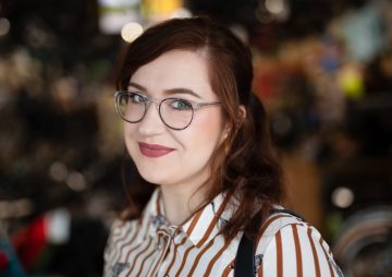 White woman with auburn hair wearing round glasses and a red and white striped shirt, background is blurred