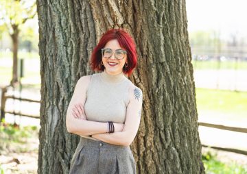 White woman with firehouse red hair, glasses, and a shoulder tattoo of willow tree branches wears a tan tank top while standing with her arms crossed in front of a large tree.