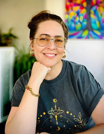 White woman with brown hair in a top bun, golden aviator glasses, and a dark great t-shirt sits in a coffee shop with a colorful painting of a tree behind her