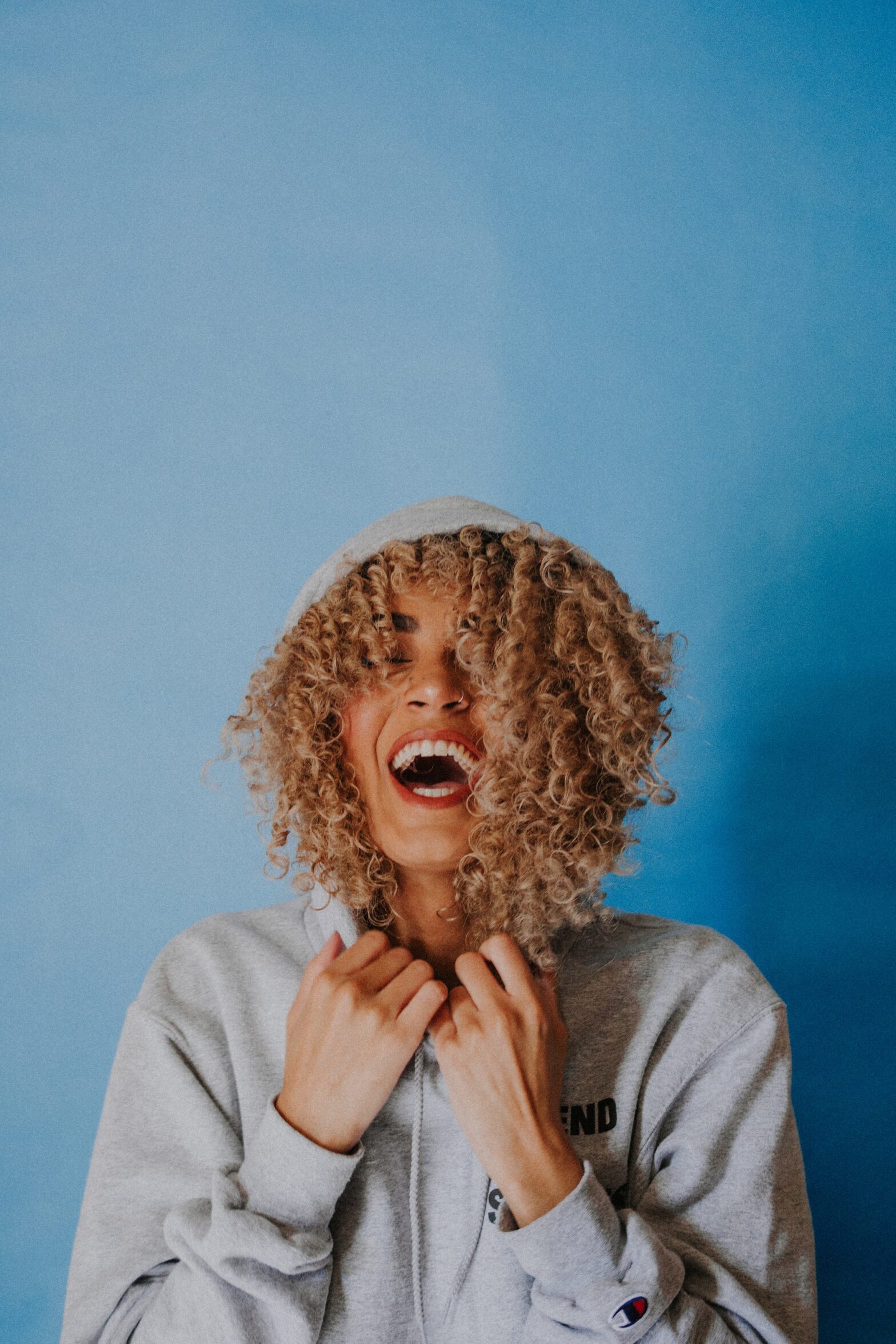 Mixed race woman with curly blond hair laughing in a sweatshirt on a blue background