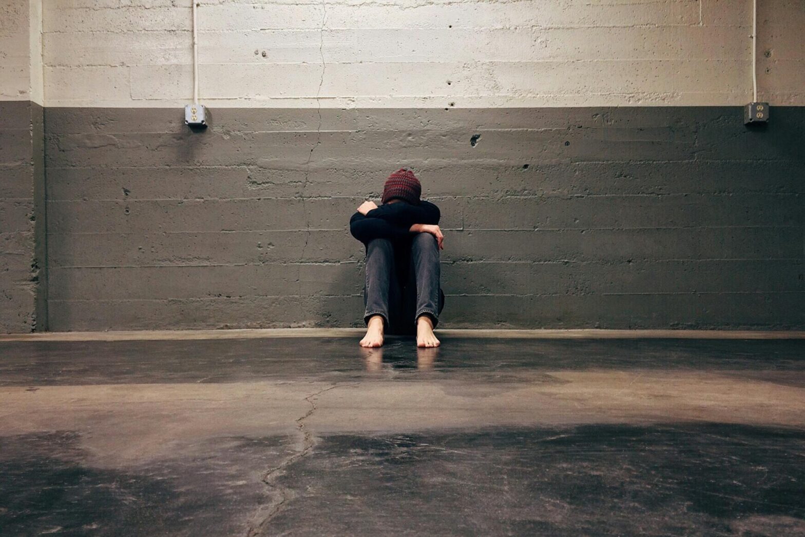 A young woman sits on the ground with her head between her knees against a grey brick wall.