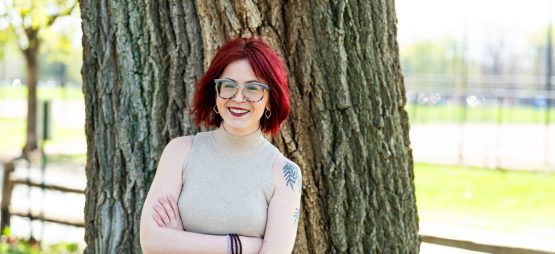 White woman with firehouse red hair, glasses, and a shoulder tattoo of willow tree branches wears a tan tank top while standing with her arms crossed in front of a large tree.