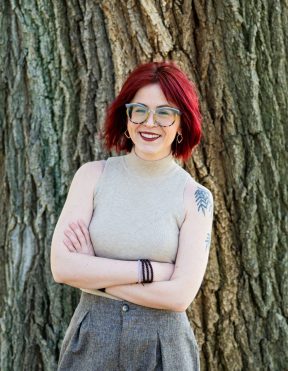 White woman with firehouse red hair, glasses, and a shoulder tattoo of willow tree branches wears a tan tank top while standing with her arms crossed in front of a large tree.