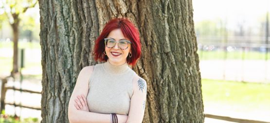 White woman with firehouse red hair, glasses, and a shoulder tattoo of willow tree branches wears a tan tank top while standing with her arms crossed in front of a large tree.