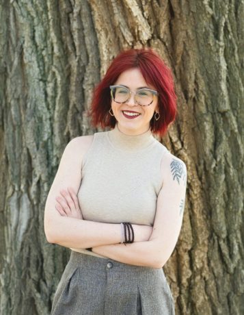 White woman with firehouse red hair, glasses, and a shoulder tattoo of willow tree branches wears a tan tank top while standing with her arms crossed in front of a large tree.