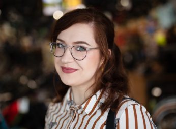 White woman with auburn hair wearing round glasses and a red and white striped shirt, background is blurred