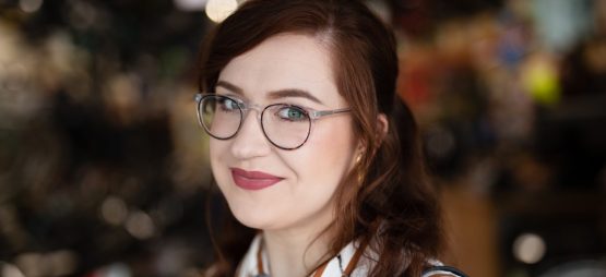 White woman with auburn hair wearing round glasses and a red and white striped shirt, background is blurred