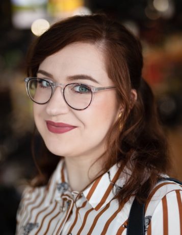 White woman with auburn hair wearing round glasses and a red and white striped shirt, background is blurred