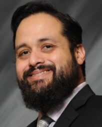 Fair skinned Hispanic man with black hair and a black beard, wearing a suit, on a grey background.