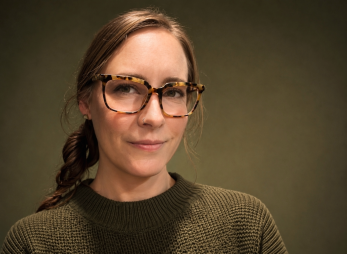 White woman with brunette hair pulled back into single braid wearing rectangular, horned, tortoiseshell glasses and an olive green sweater. Background is portrait-style solid color olive green.
