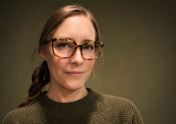 White woman with brunette hair pulled back into single braid wearing rectangular, horned, tortoiseshell glasses and an olive green sweater. Background is portrait-style solid color olive green.