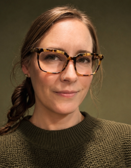 White woman with brunette hair pulled back into single braid wearing rectangular, horned, tortoiseshell glasses and an olive green sweater. Background is portrait-style solid color olive green.