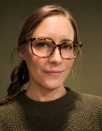 White woman with brunette hair pulled back into single braid wearing rectangular, horned, tortoiseshell glasses and an olive green sweater. Background is portrait-style solid color olive green.