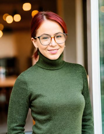 White woman with glasses and red hair which is pulled back into a short pony tail. She wears a green turtleneck sweater.