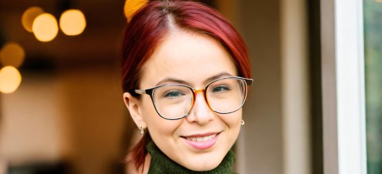 White woman with glasses and red hair which is pulled back into a short pony tail. She wears a green turtleneck sweater.