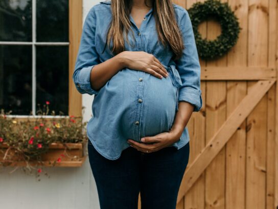 A pregnant woman stands in front of a window and a wooden door, she is wearing black pants and a blue jean shirt, she is holding her belly and her face is cut out of frame