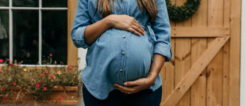 A pregnant woman stands in front of a window and a wooden door, she is wearing black pants and a blue jean shirt, she is holding her belly and her face is cut out of frame