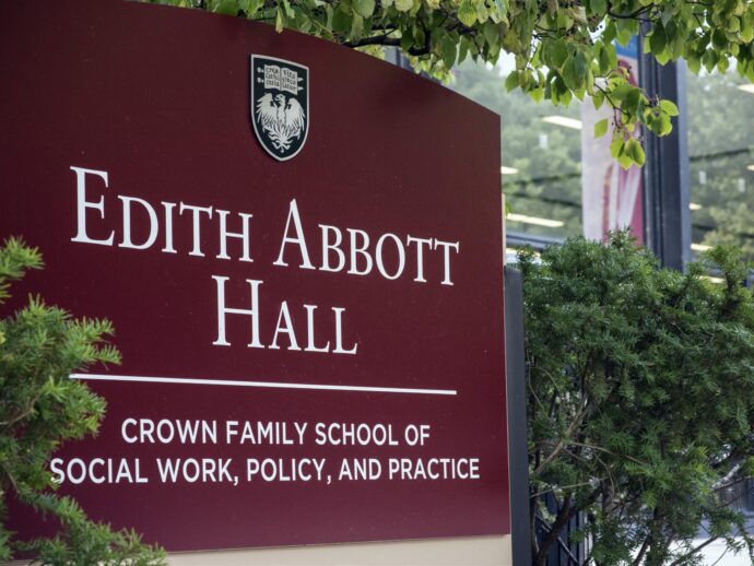 An exterior photo of the Edith Abbott Hall at the Crown School of Social Work at University of Chicago. A maroon sign labeling the building against leafy green foliage and the glass building.