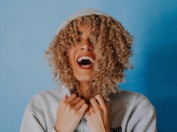 Mixed race woman with curly blond hair laughing in a sweatshirt on a blue background