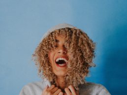 Mixed race woman with curly blond hair laughing in a sweatshirt on a blue background