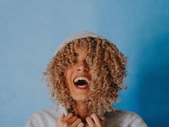 Mixed race woman with curly blond hair laughing in a sweatshirt on a blue background