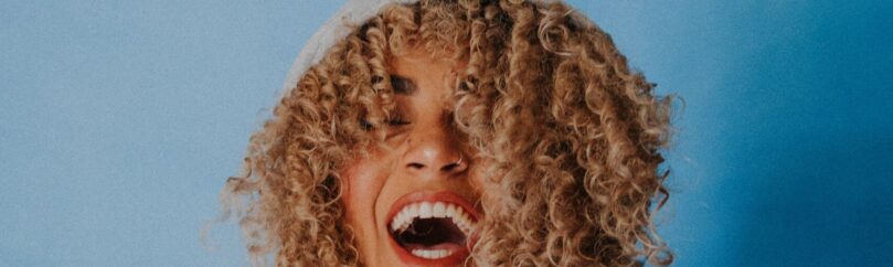 Mixed race woman with curly blond hair laughing in a sweatshirt on a blue background
