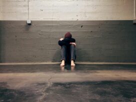 A young woman sits on the ground with her head between her knees against a grey brick wall.