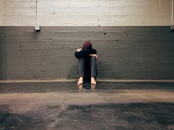 A young woman sits on the ground with her head between her knees against a grey brick wall.