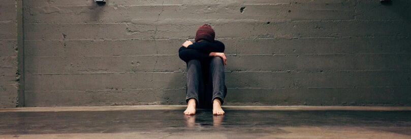 A young woman sits on the ground with her head between her knees against a grey brick wall.